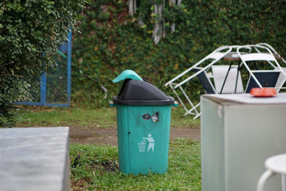 Multiple purple garden waste bags, tied with red string, are stacked along the edge of a mown grassy lawn. The background features dense, well-maintained green hedges and trees, with a glimpse of resi